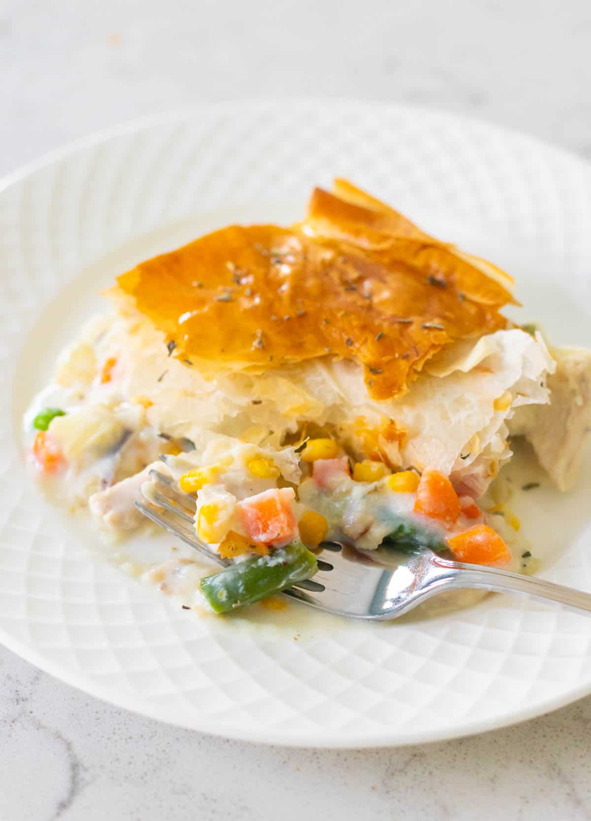 The square of chicken pot pie is on a white plate. The vegetables are spilling out from the filling so you can see the ingredients included.