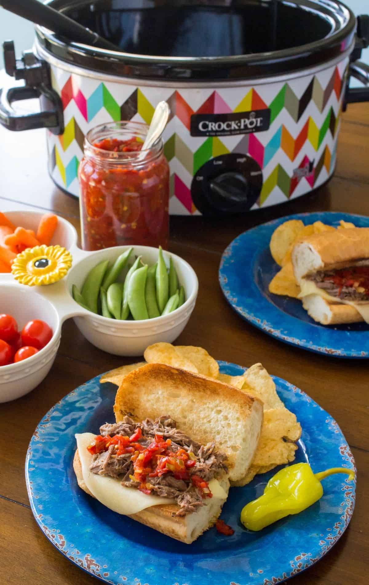 The plated italian beef sandwiches are on a table next to a raw veggie platter and a Crockpot in the background.