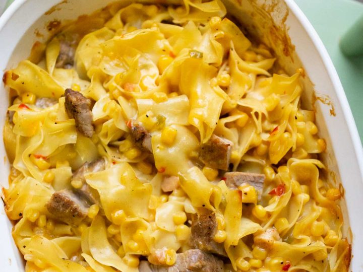 A white baking dish is filled with wavy egg noodles, chopped pork, and corn and red peppers to show the final baked saucy pork and noodle bake casserole.