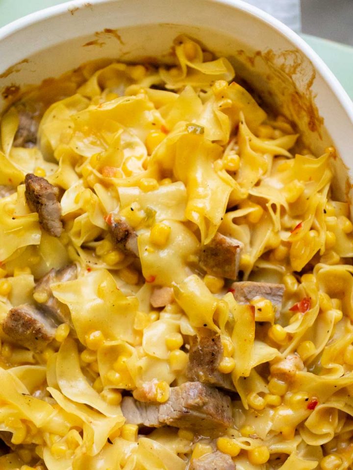 A white baking dish is filled with wavy egg noodles, chopped pork, and corn and red peppers to show the final baked saucy pork and noodle bake casserole.