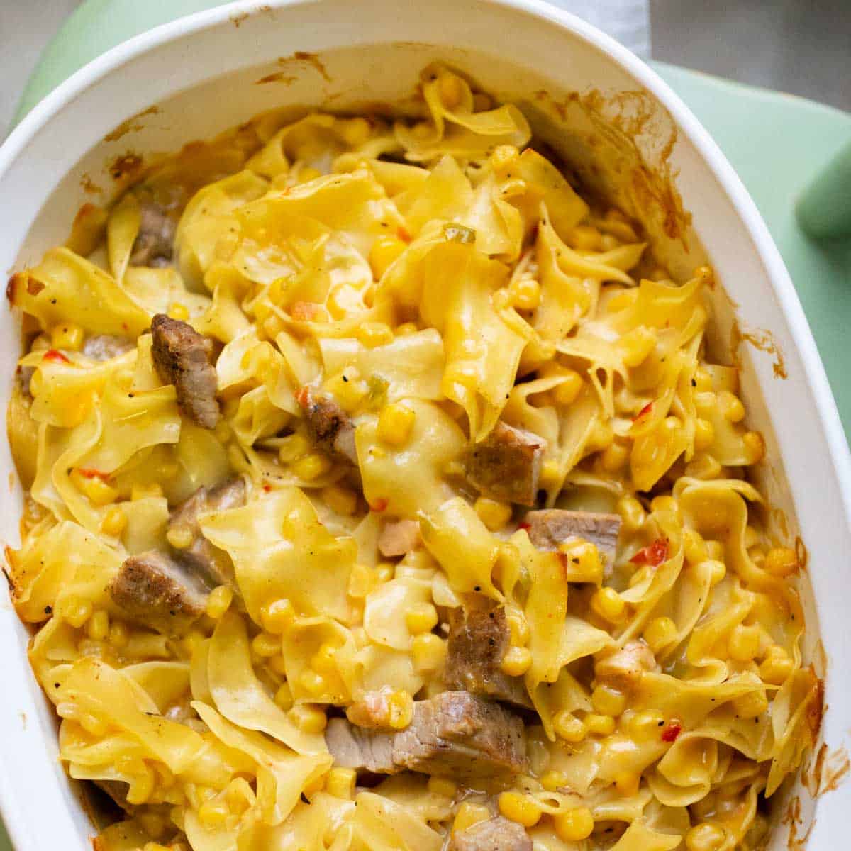 A white baking dish is filled with wavy egg noodles, chopped pork, and corn and red peppers to show the final baked saucy pork and noodle bake casserole.