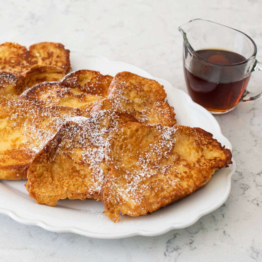 Crispy french toast slices on a platter sprinkled with powdered sugar and a cup of maple syrup on the side.