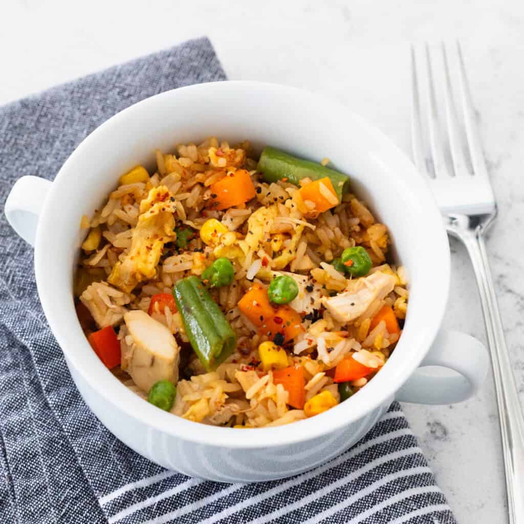 A small white bowl is filled with chicken fried rice and veggies. A fork rests on the side.