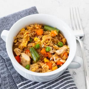 A small white bowl is filled with chicken fried rice and veggies. A fork rests on the side.