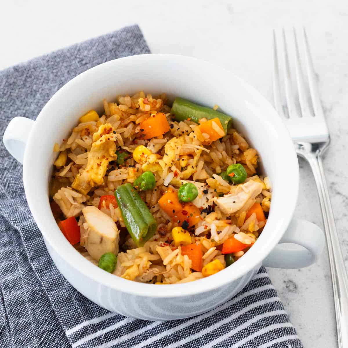 A small white bowl is filled with chicken fried rice and veggies. A fork rests on the side.
