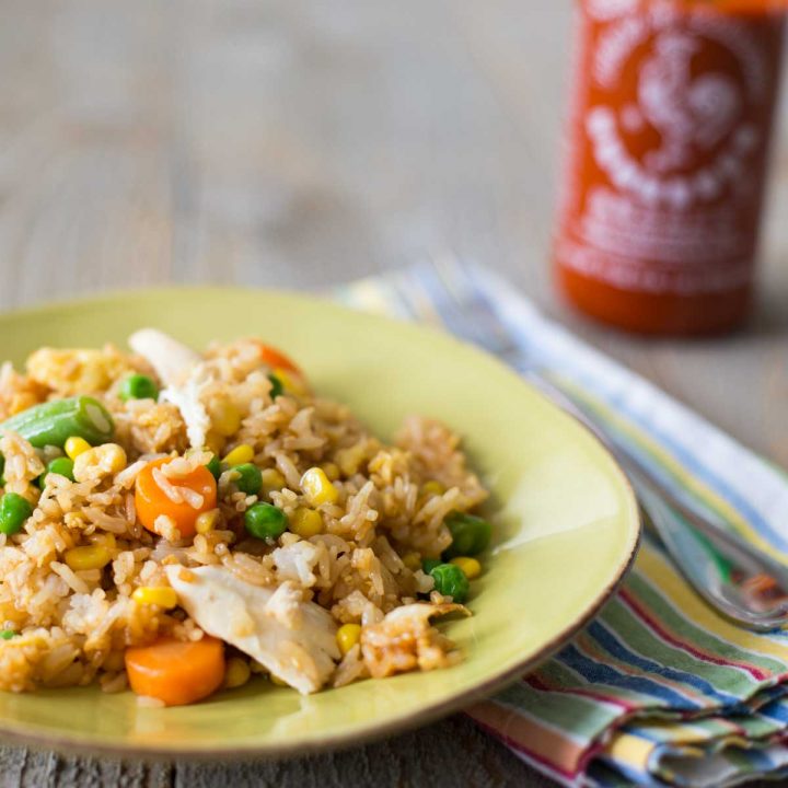 A plate of chicken fried rice with a bottle of sriracha sauce in the background.
