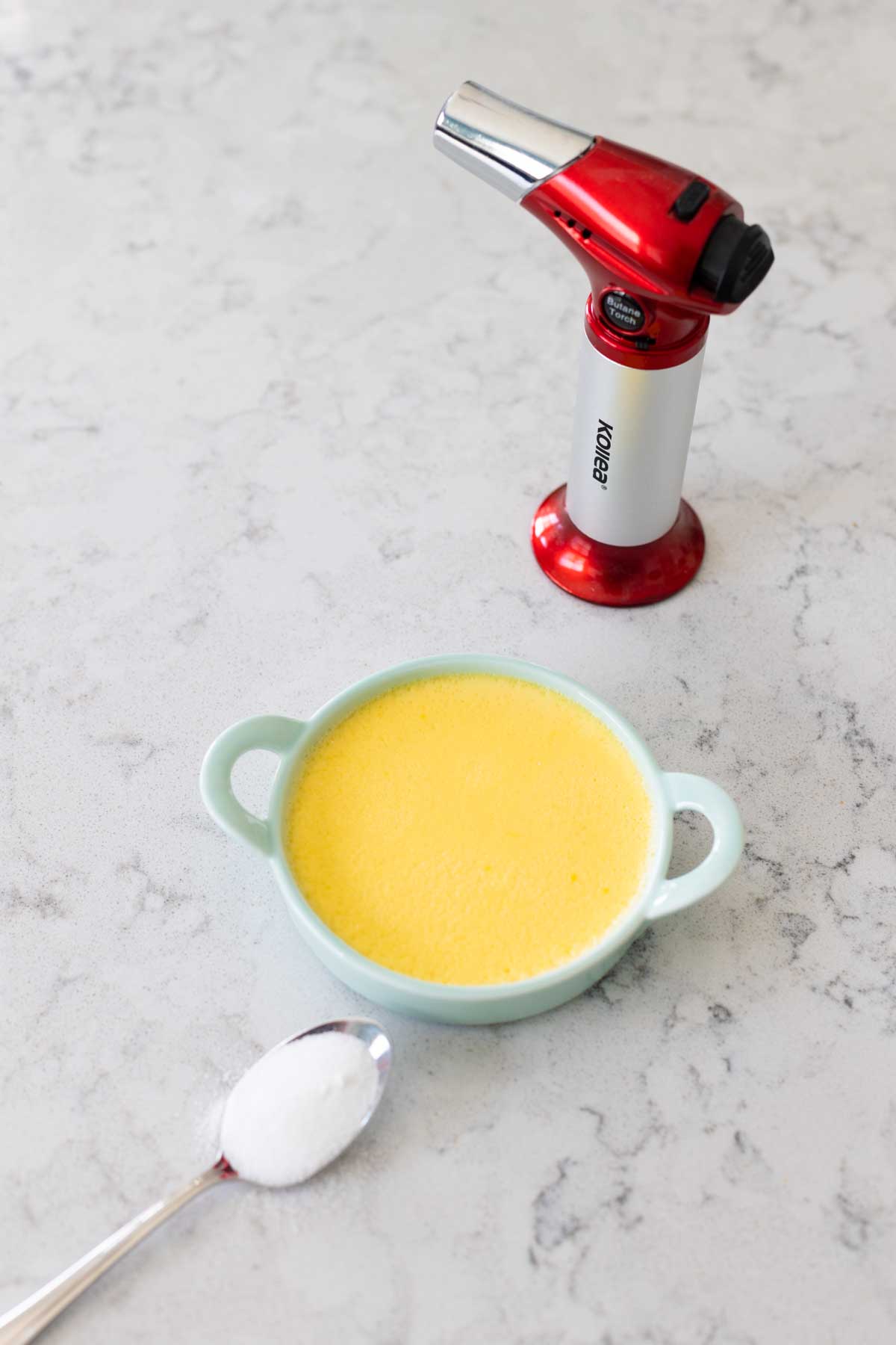 A kitchen torch, a spoon of sugar, and one Crème Brûlée ramekin.