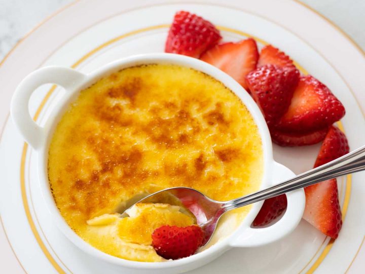 A serving of creme brulee with fresh strawberries. A spoon has cracked through the sugar topping.