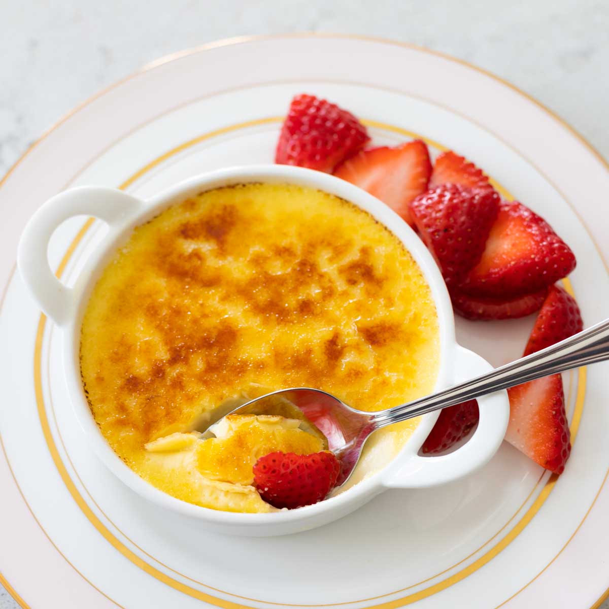 A serving of creme brulee with fresh strawberries. A spoon has cracked through the sugar topping.