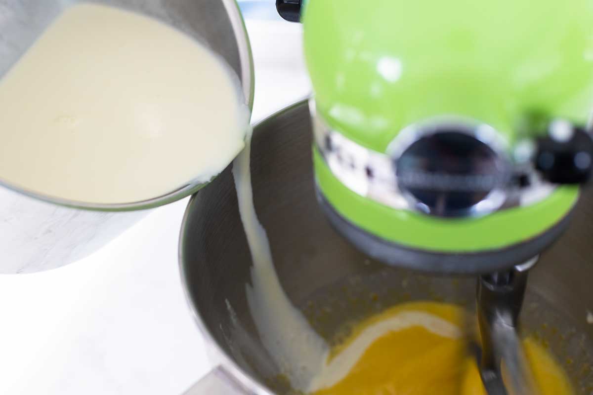 The milk is being poured down the inside side of the mixing bowl while the paddle attachment is turning to keep the eggs moving.