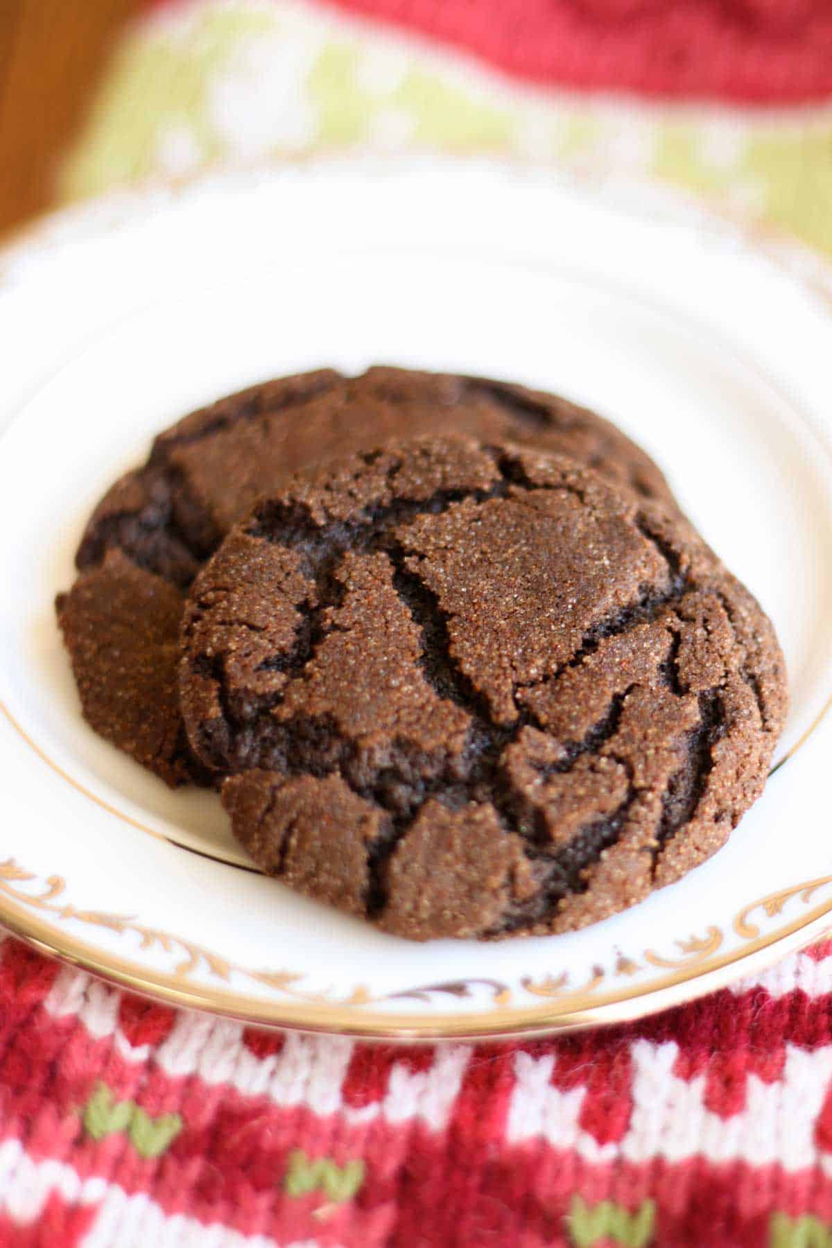 Two chocolate cookies with crinkles over the surface sit on a china plate. A knitted stocking sits under the plate.