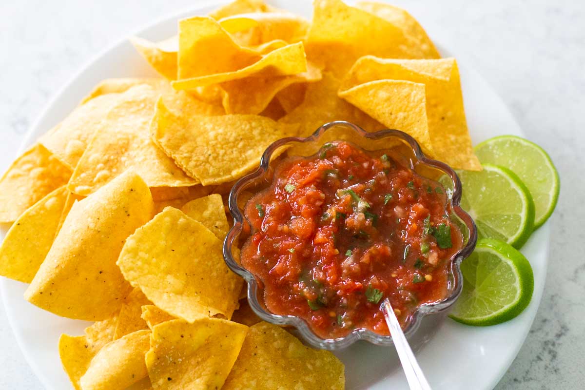 A bowl of the finished salsa is in a bowl with a pile of tortilla chips and a few slices of lime.