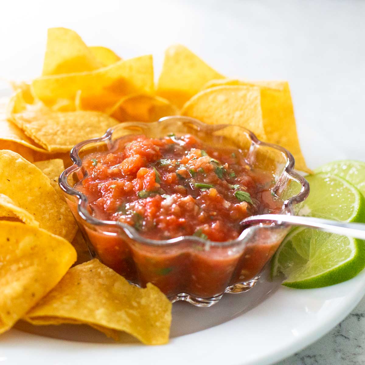 A bowl of homemade salsa has a spoon on the right and a pile of restaurant style tortilla chips on the plate around it.