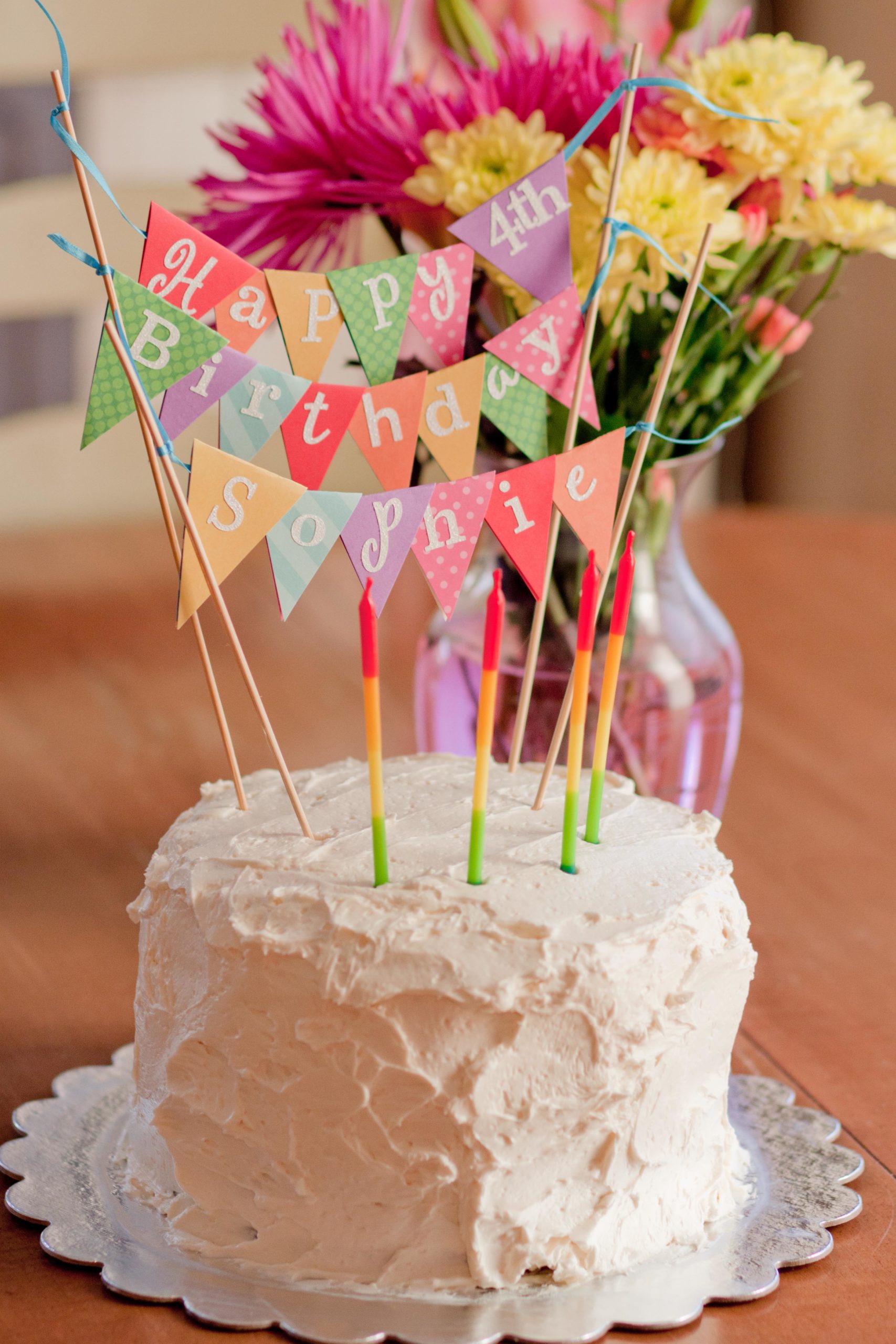 A birthday cake has a bunting cake topper that says "Happy 4th Birthday" in rainbow flags.