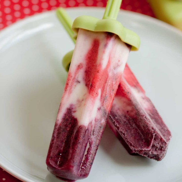 Red, white, and blue firecracker popsicles are stacked on a plate. They have a green plastic handle.