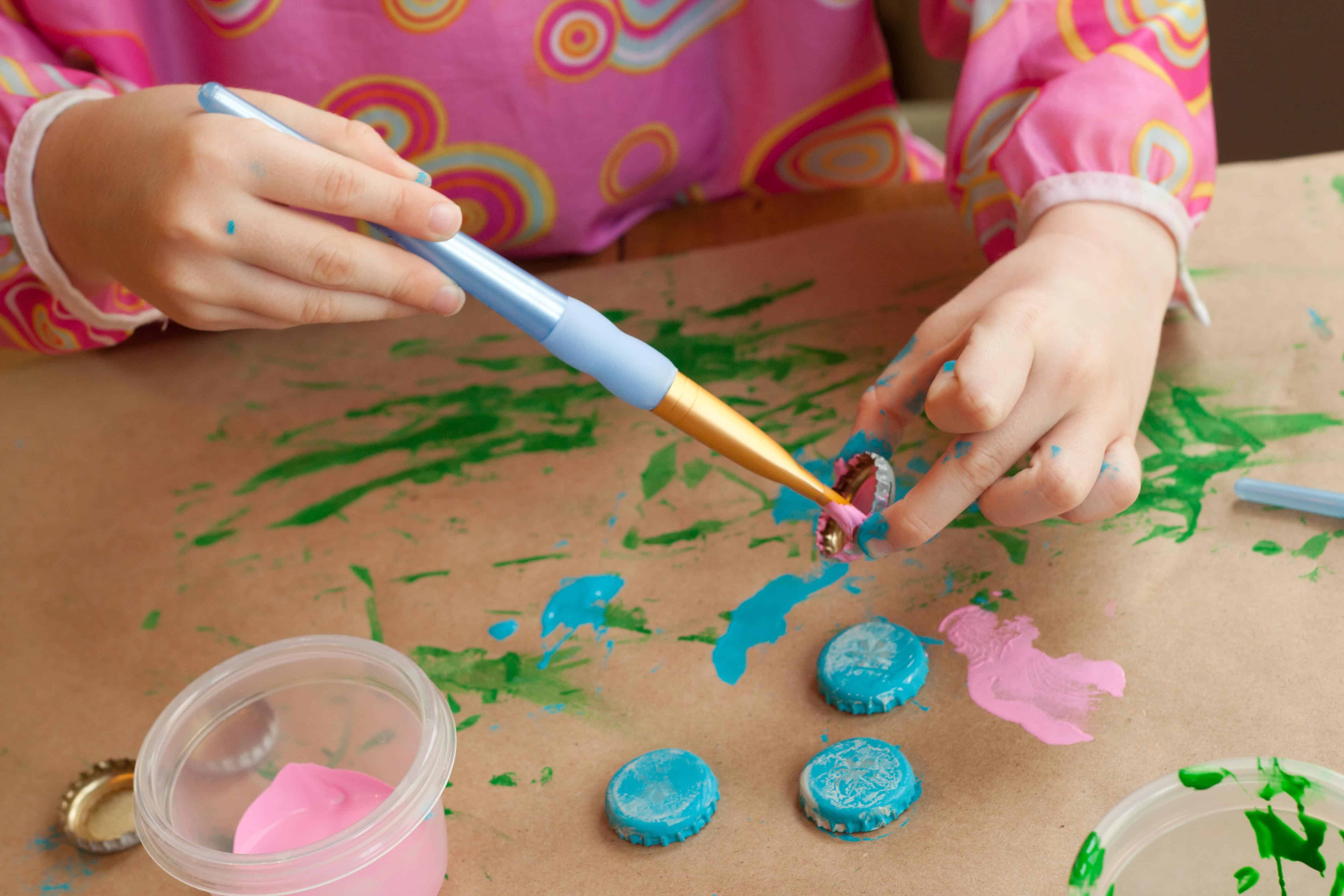 A child paints recycled bottle caps with pink and blue paint to create colorful garden markers on a craft-covered table.