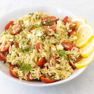 A white bowl is filled with the lemon orzo pasta salad. Tomatoes and dill are sprinkled throughout.