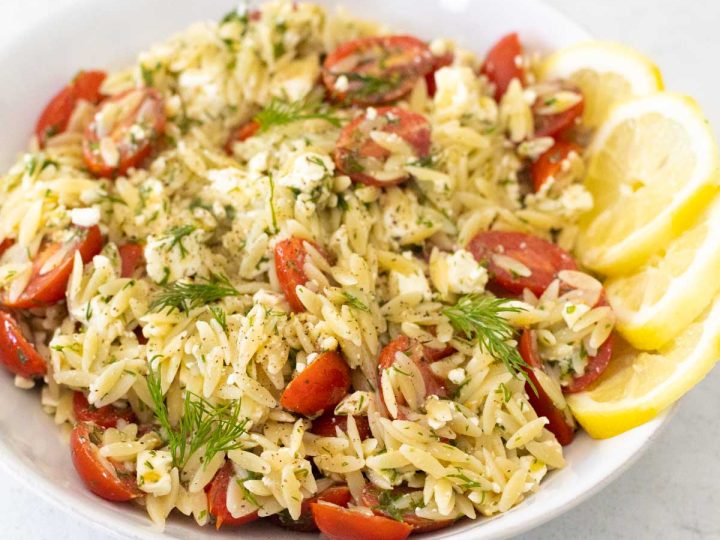 A white bowl is filled with the lemon orzo pasta salad. Tomatoes and dill are sprinkled throughout.