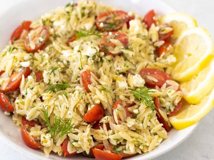 A white bowl is filled with the lemon orzo pasta salad. Tomatoes and dill are sprinkled throughout.