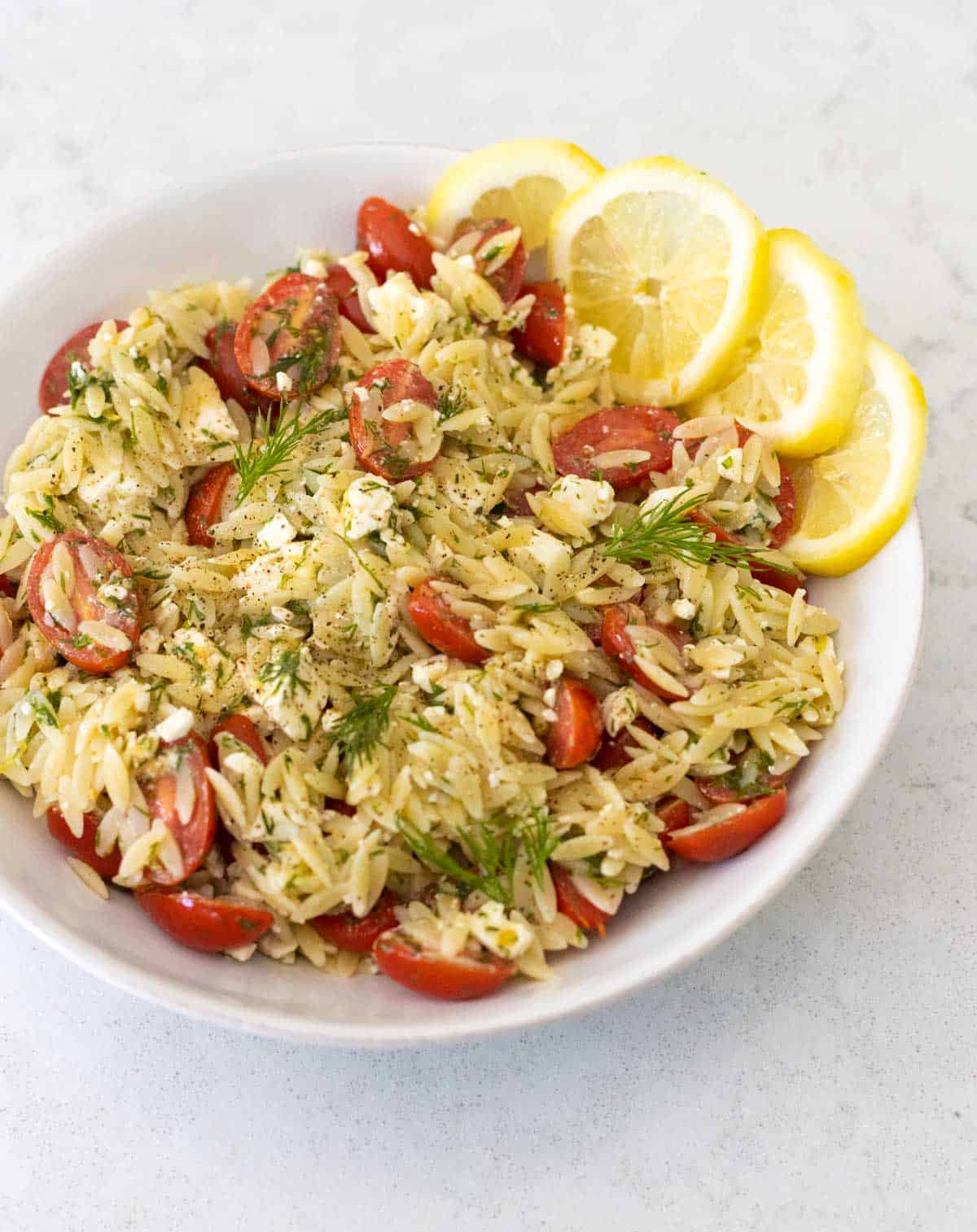 A white bowl is filled with the lemon orzo pasta salad. Tomatoes and dill are sprinkled throughout.