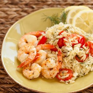 A plate of orzo pasta with fresh tomatoes sprinkled with fresh dill has roasted shrimp and lemon slices in the back.