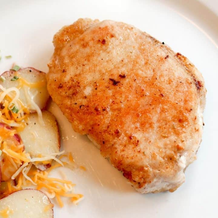 A cooked, pan-fried pork chop sits on a dinner plate next to potatoes.