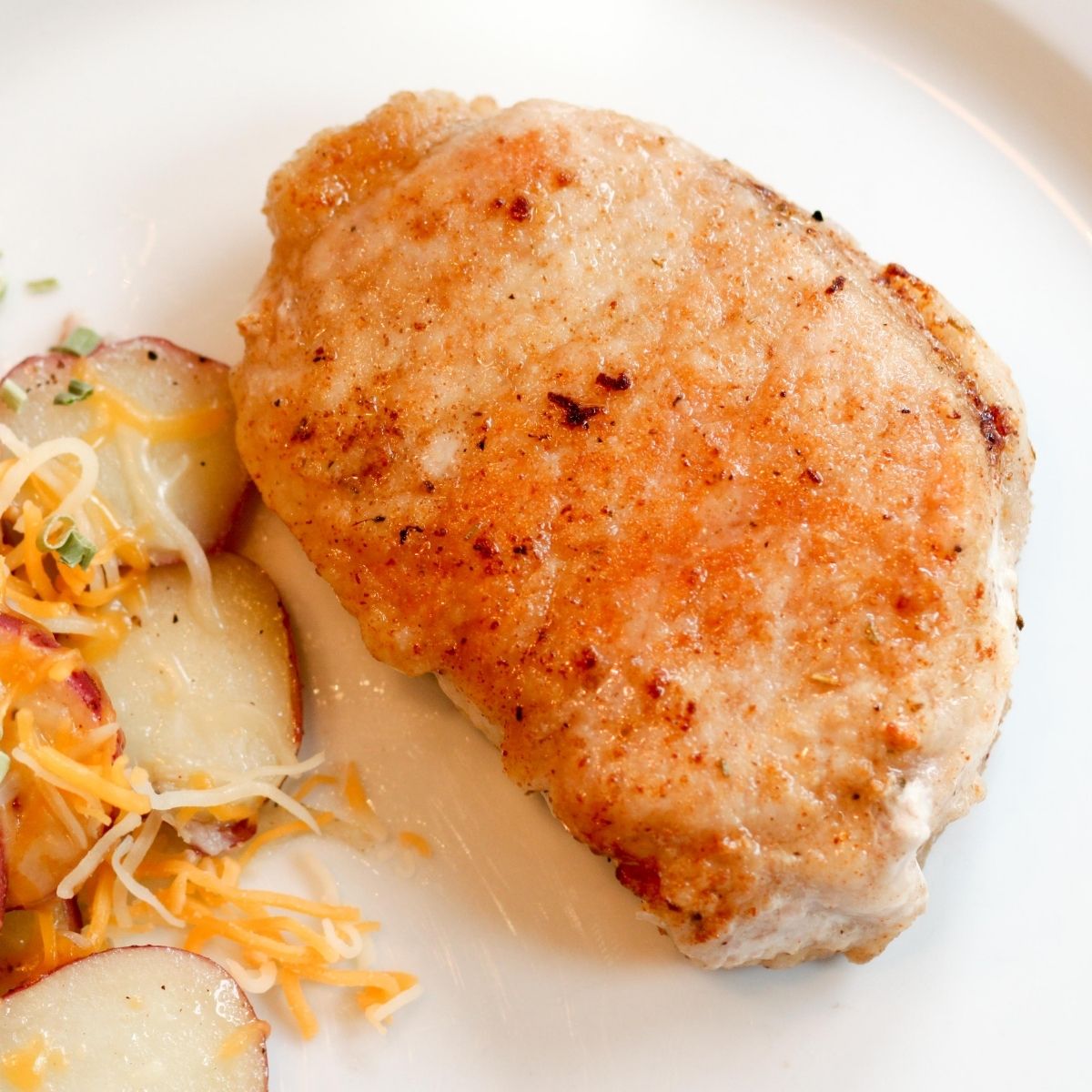 A cooked, pan-fried pork chop sits on a dinner plate next to potatoes.