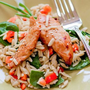 A plate of orzo. pasta with two chicken tenderloins on top. Fresh spinach and chopped veggies are mixed in.