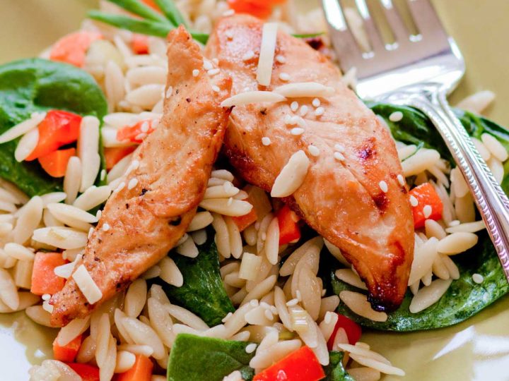 A plate of orzo. pasta with two chicken tenderloins on top. Fresh spinach and chopped veggies are mixed in.