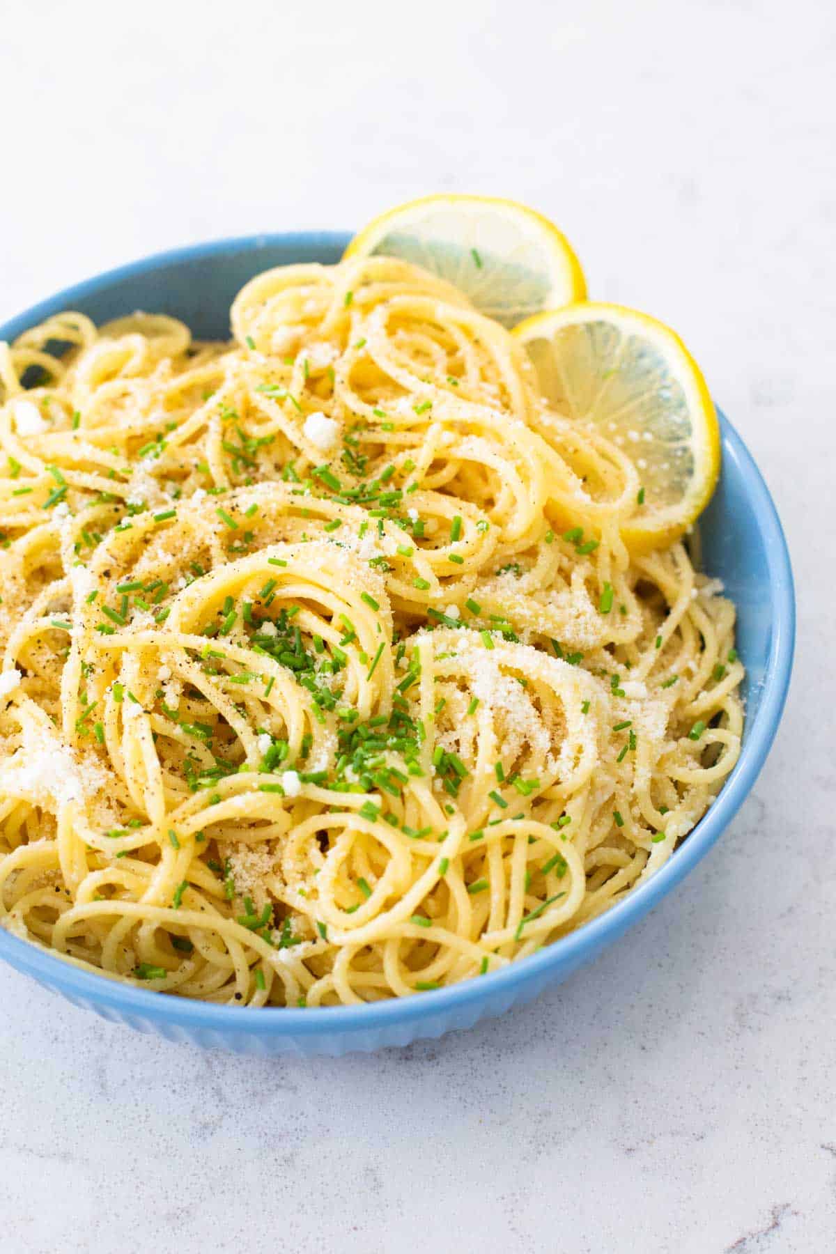 A bowl of spaghetti in a lemon butter sauce has fresh chives and slices of fresh lemon on the side.