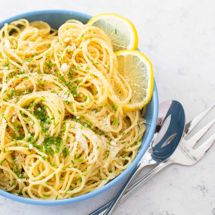 A bowl of spaghetti in a lemon butter sauce has fresh chives and slices of fresh lemon on the side.