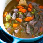 A blue dutch oven has a wooden spoon stirring beef stew.
