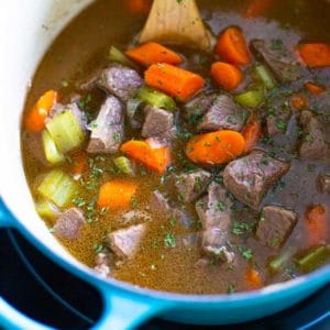 A blue dutch oven has a wooden spoon stirring beef stew.