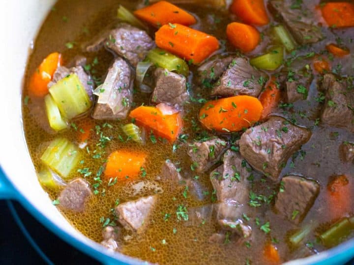 A blue dutch oven has a wooden spoon stirring beef stew.