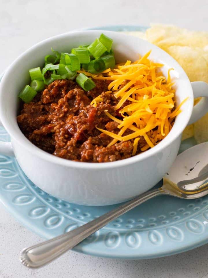 A white soup bowl filled with thick ground beef chili topped with shredded cheese and sliced green onions. There's a spoon and a pile of potato chips on the side.
