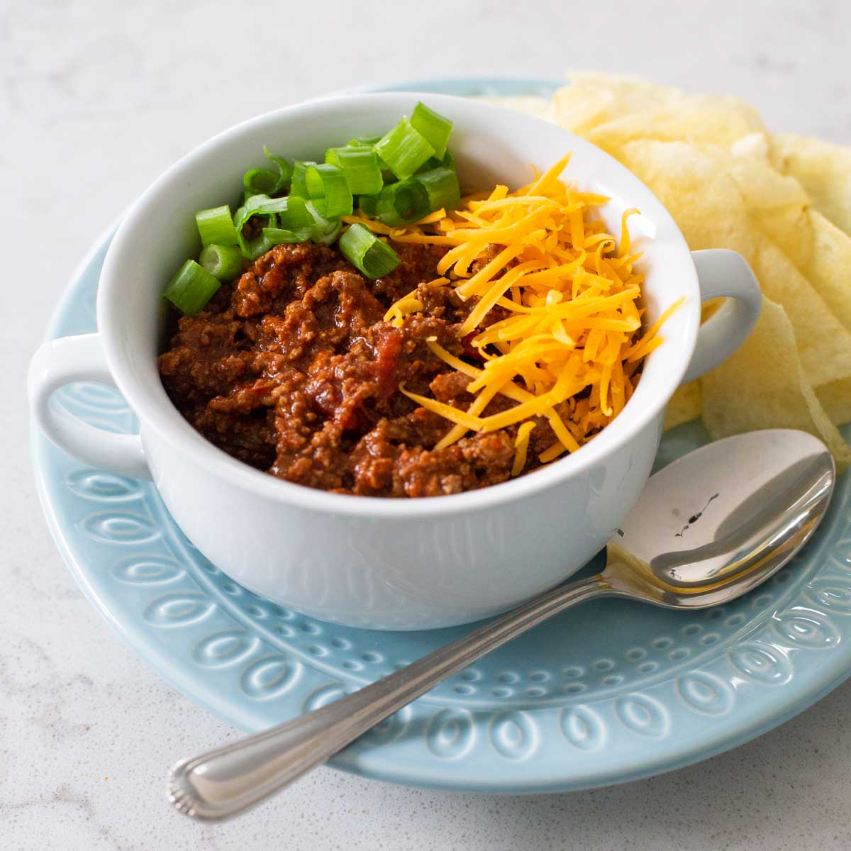 A white soup bowl filled with thick ground beef chili topped with shredded cheese and sliced green onions. There's a spoon and a pile of potato chips on the side.