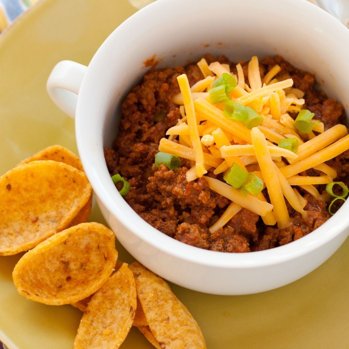 A bowl of very thick chili with shredded cheese and green onions on top and Frito chips on the side for dunking.