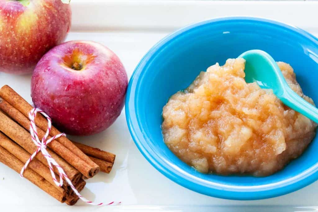 A bowl of applesauce sits next to fresh apples and a bundle of cinnamon sticks.