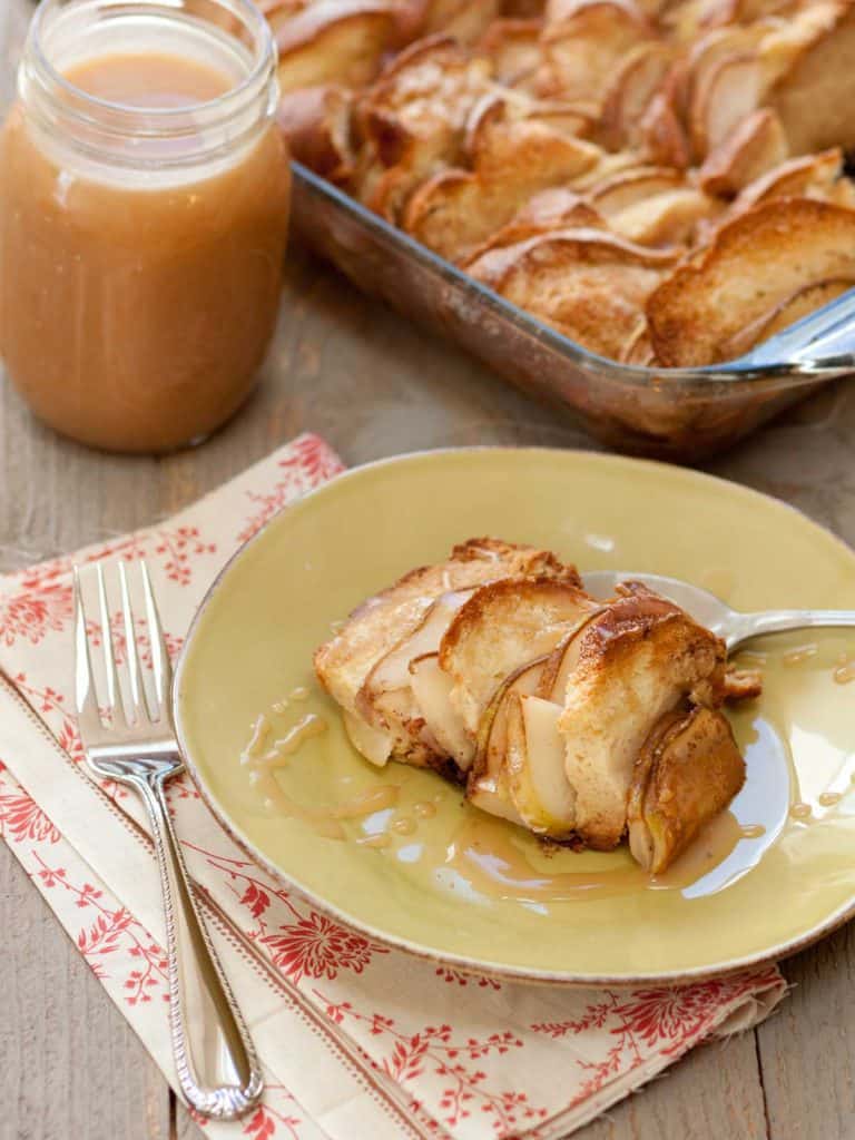 A serving of bread pudding with sliced pears sits on a plate. A jar of salted caramel sauce sits in the background next to the pan of bread pudding.