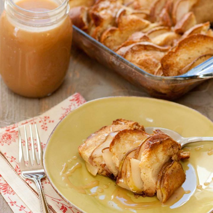 A serving of bread pudding with sliced pears sits on a plate. A jar of salted caramel sauce sits in the background next to the pan of bread pudding.