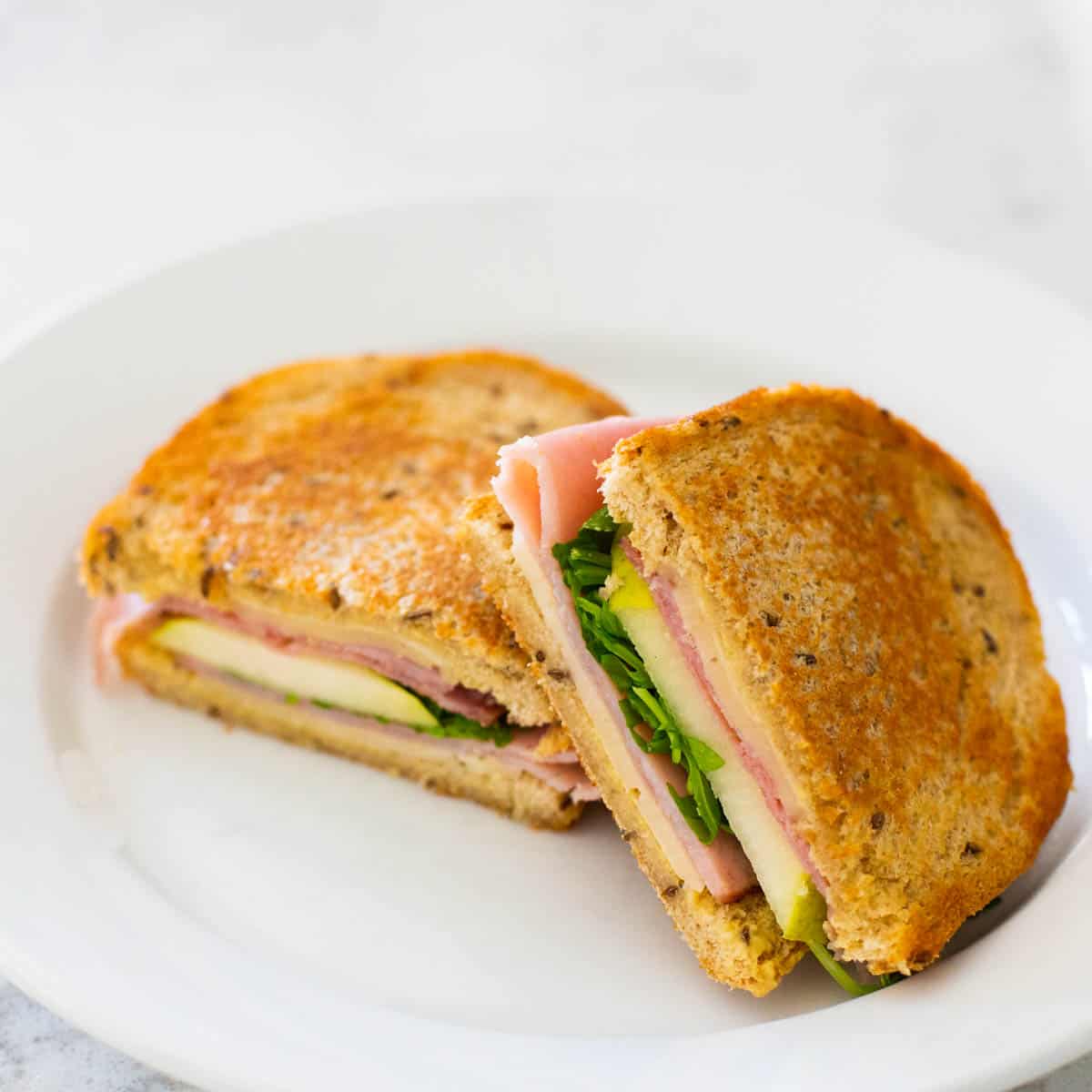 The finished grilled sandwich is on a white plate. You can see the layers of arugula, fresh pear slices, and ham and havarti cheese.