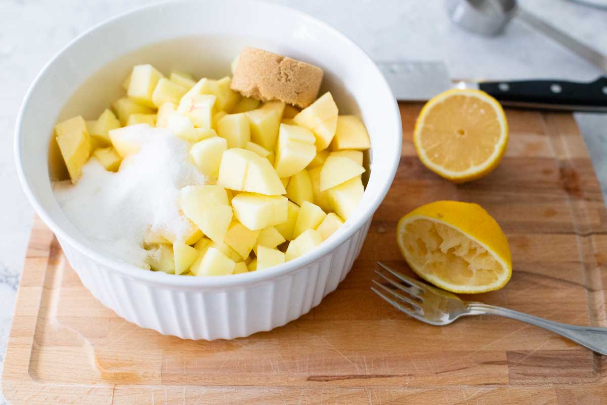 A microwave container is filled with the chopped apples, sugars, and has a fresh squeezed lemon on the board next to it.