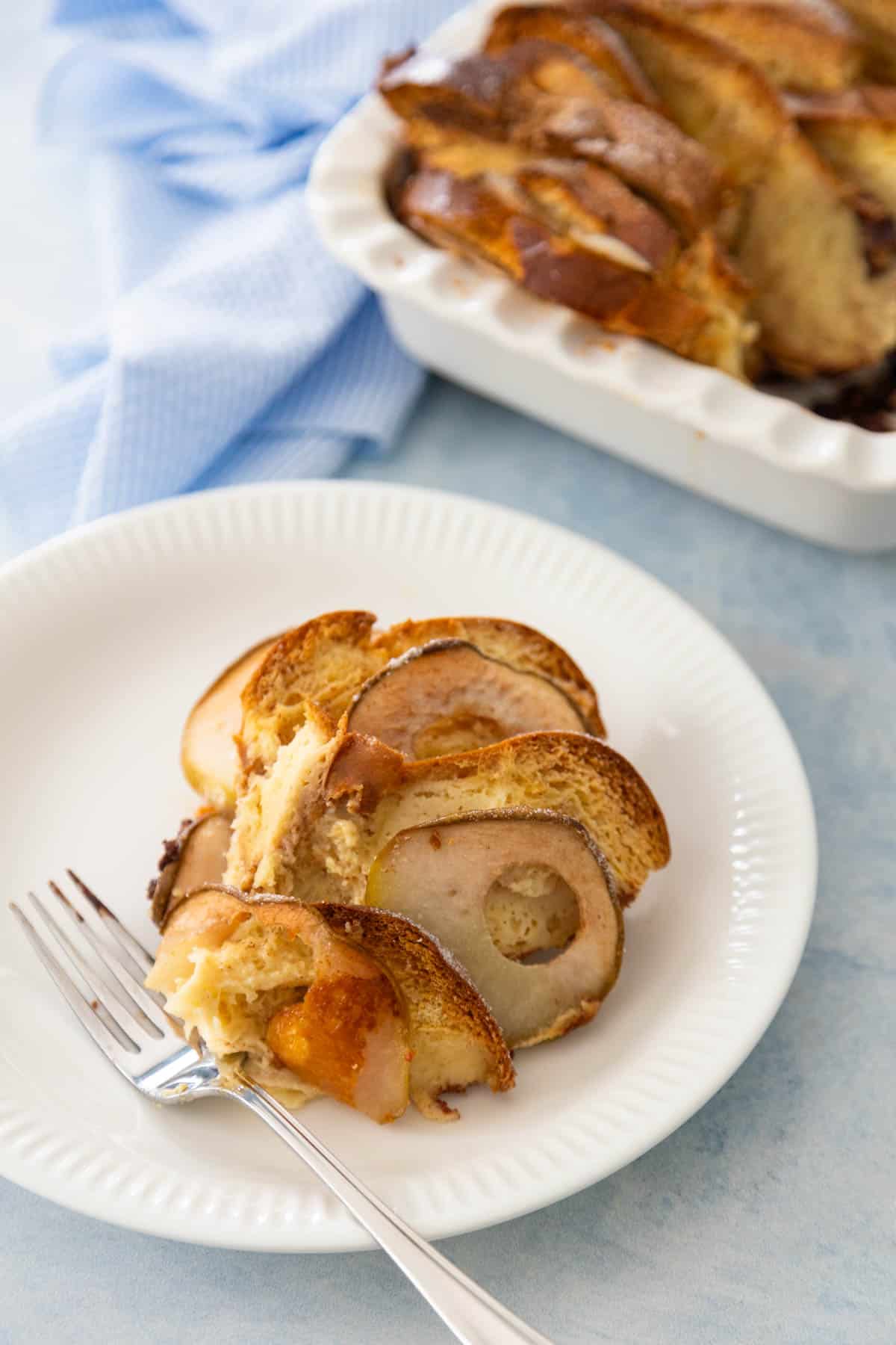 The serving of pear bread pudding is on a white plate next to the baking dish of dessert.