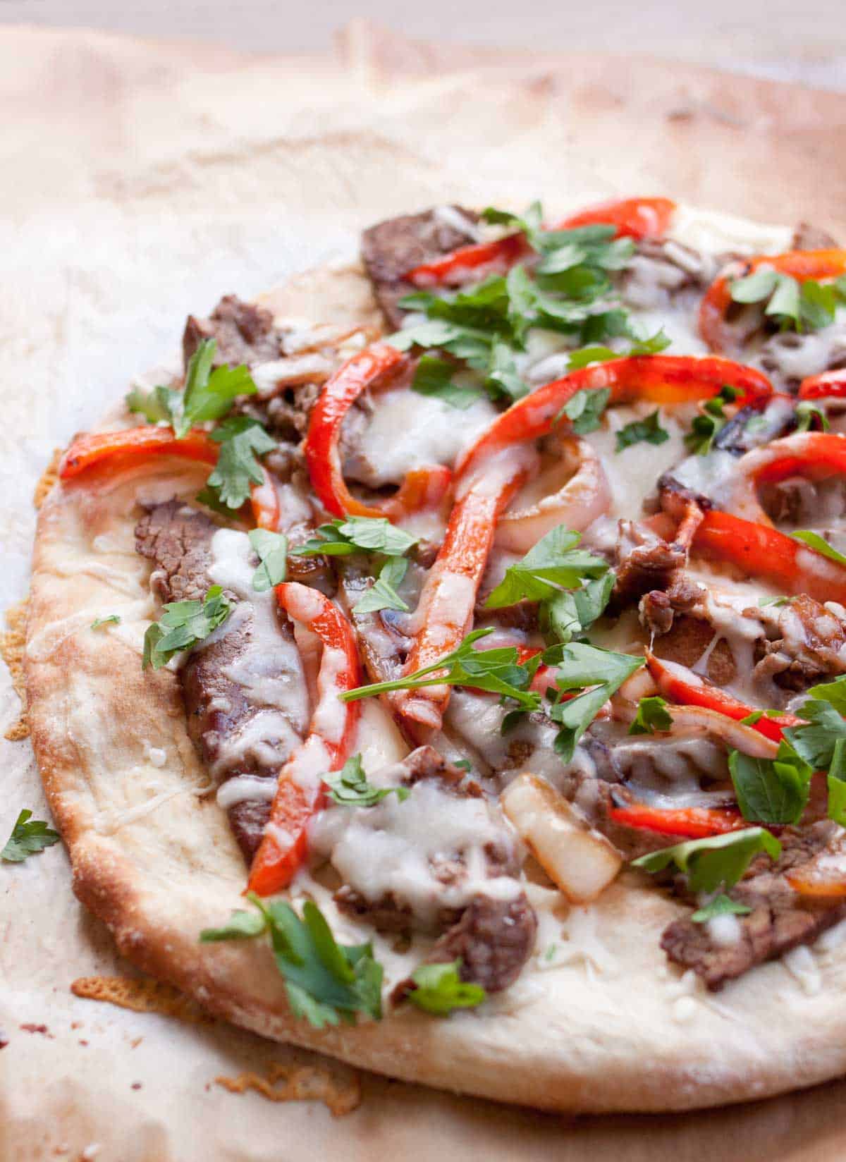 A flatbread pizza with strips of steak and red peppers is covered in melted cheese and fresh parsley.