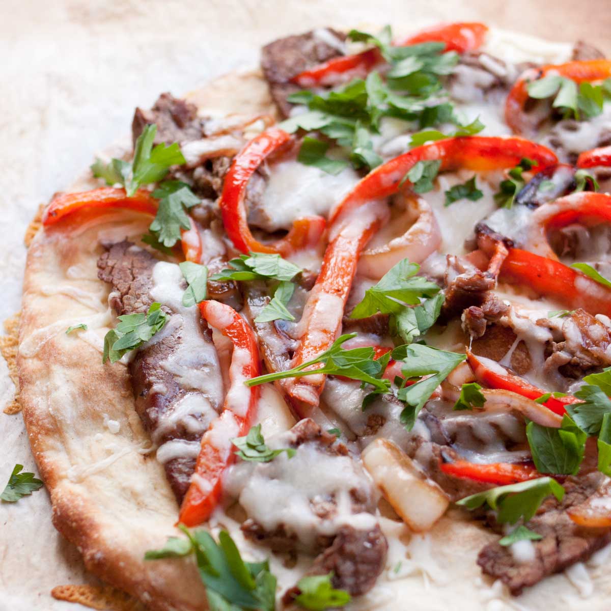 A flatbread pizza with strips of steak and red peppers is covered in melted cheese and fresh parsley.