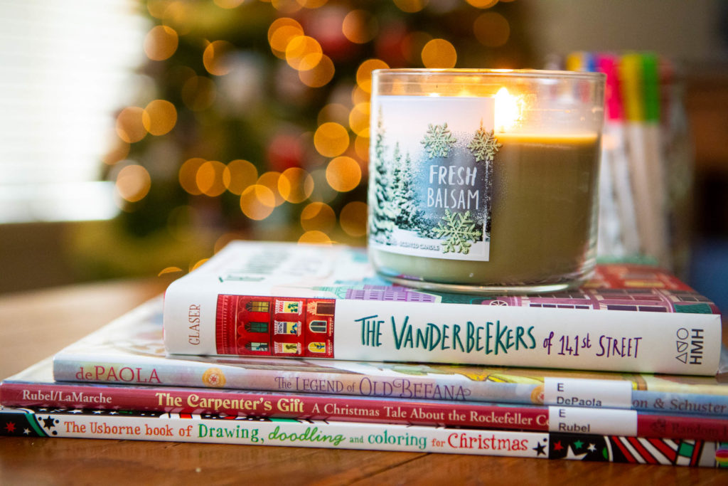 A stack of Christmas books sit under a lit candle in front of a Christmas tree.