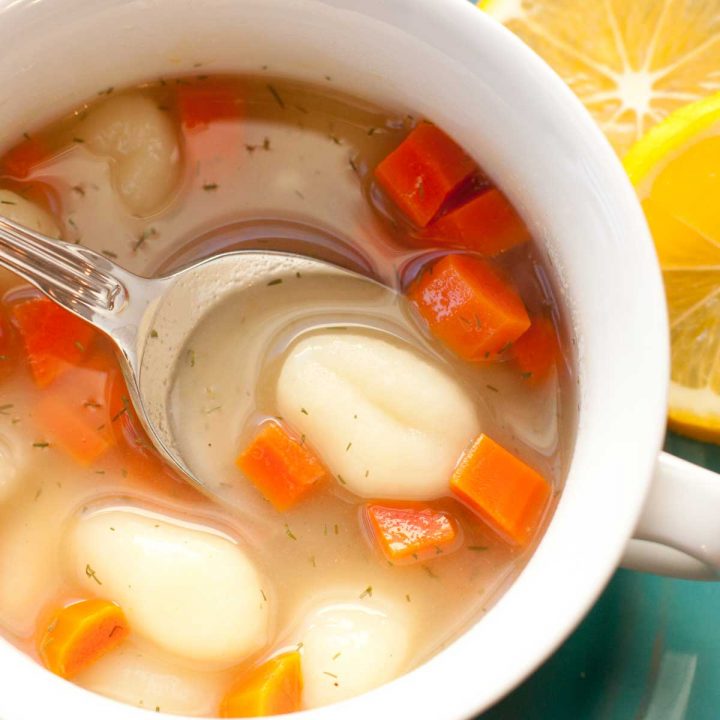 A bowl of gnocchi soup has chunks of diced carrot and a clear broth. Slices of lemon are on the plate next to the bowl.