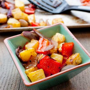 A serving bowl of roasted pineapple and pepper chunks sits in front of a roasting pan with spatula.