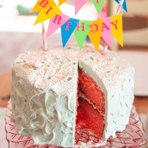 A slice is missing from a pink strawberry layer cake with aqua-tinted frosting. There are pastel sprinkles and a birthday pennant decorating the top of the cake.