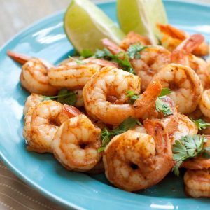 A blue plate has a pile of spicy shrimp with two lime wedges and a sprinkle of fresh cilantro over the top.